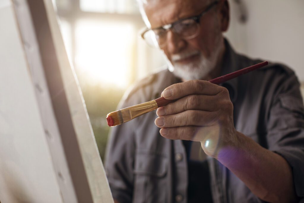 An older man holds a paintbrush in his hand a few inches away from a canvas as he contemplates what to paint