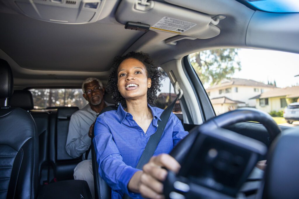 A woman driving a vehicle looks in her rearview mirror while speaking with a Medicare beneficiary sitting in the backseat